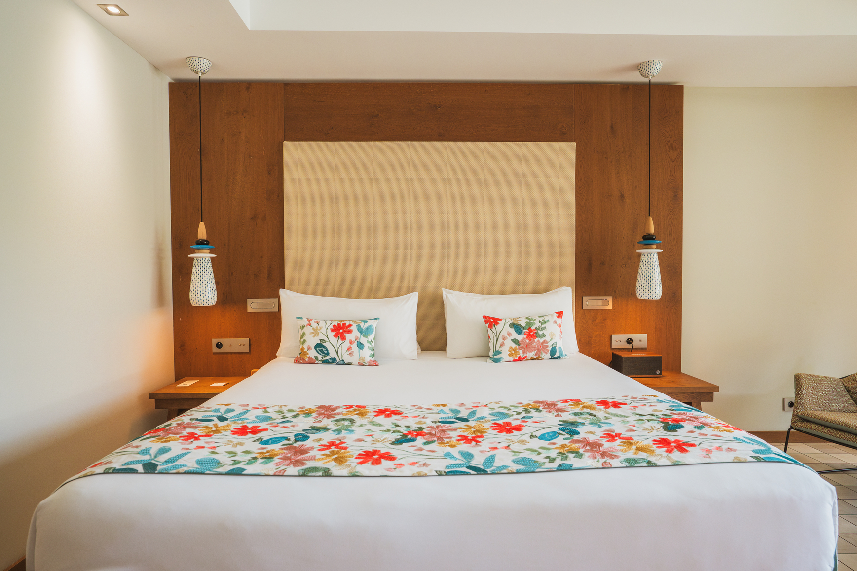 Chambre Deluxe Wood avec tête de lit monumentale en bois massif, jeté de lit floral coloré et ambiance lumineuse chaleureuse.